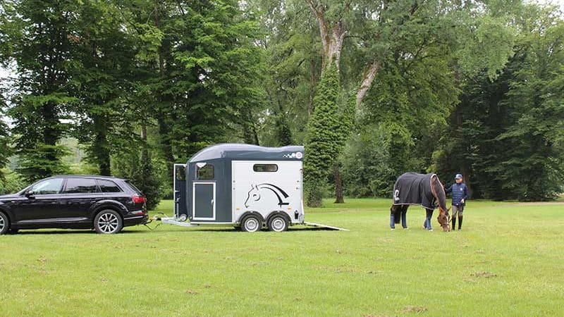 Una furgoneta enganchada a un coche en un prado verde. Un caballo y un jinete pastan cerca.