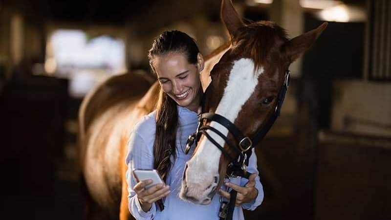 Mujer joven mirando su smartphone y sonriendo. Su caballo le mira por encima del hombro.