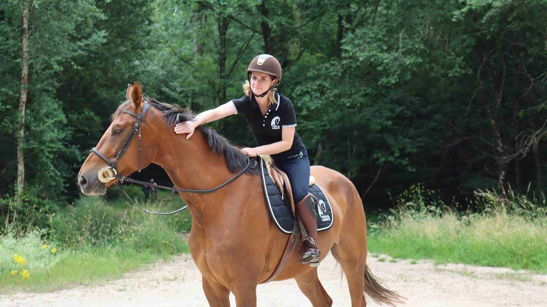 Una jinete inmóvil acaricia a su caballo en el bosque, sonriendo.